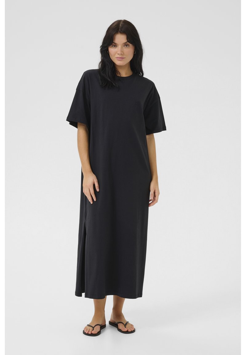 Czarna, sięgająca do kolan sukienka T-shirt z miękkiego materiału, z krótkimi rękawami, bocznymi rozcięciami i luźnym krojem. Noszona z czarnymi klapkami.