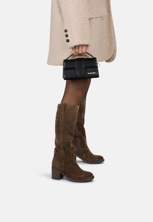 Femme portant des bottes en daim marron hautes jusqu'au genou et un manteau en laine beige, tenant un petit sac Jacquemus noir par sa poignée.