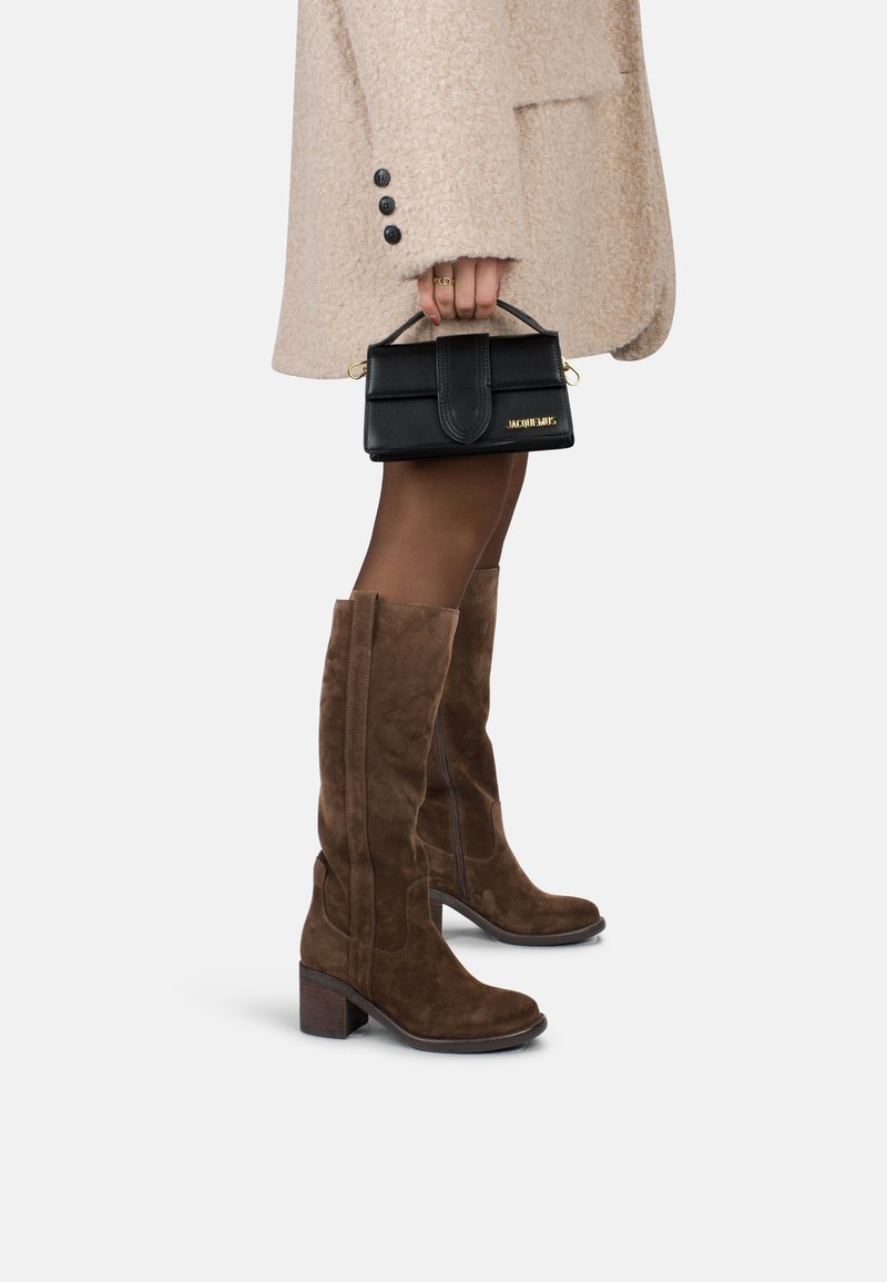 Femme portant des bottes en daim marron hautes jusqu'au genou et un manteau en laine beige, tenant un petit sac Jacquemus noir par sa poignée.