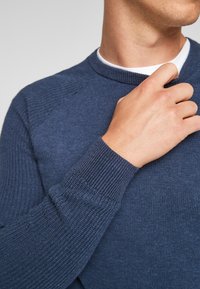 Dunkelblauer Strickpullover mit gerippten Bündchen und Kragen, ausgestattet mit einem strukturierten Muster und Raglanärmeln. Hand in der Nähe des Kragens positioniert.