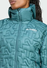 Teal gewatteerde jas met een hoge kraag, ritssluiting en een klein wit logo. De stof heeft een gladde textuur met geometrische patronen.