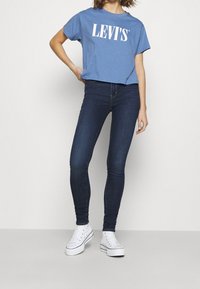 Mujer vestida con una camiseta recortada azul de Levi's, jeans ajustados azul oscuro y zapatillas altas blancas, de pie con una mano en la cadera.