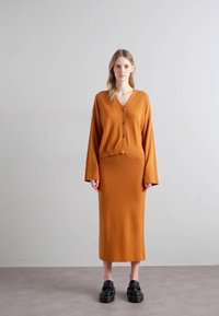 Cardigan en tricot orange brûlé à manches longues et cinq boutons, associé à une jupe midi moulante assortie. Chaussures à plateforme noires.