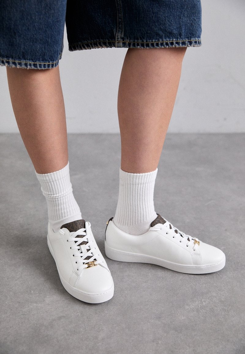 Personne portant des baskets blanches avec des détails dorés, des chaussettes blanches côtelées et un pantalon en jean court, debout sur un sol gris.