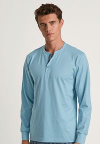 Lichtblauwe langslievende top met een henley halslijn, voorzien van twee knopen en een rechte zoom. Glad materiaal en aansluitende manchetten.