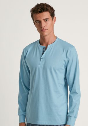 Haut à manches longues bleu clair avec un col henley, présentant deux boutons et un ourlet droit. Tissu lisse et poignets ajustés.