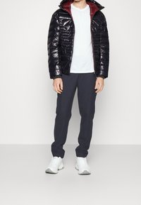 Polstrovaná černá bunda s lesklým povrchem, červenou podšívkou, na zip, nošená přes bílou košili a tmavé kalhoty, doplněná o bílé tenisky.