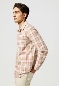 Camisa de cuadros de manga larga en beige con cuadros rojos. Cuenta con un collar con botones y un bolsillo en el pecho. Se lleva con pantalones de color claro.