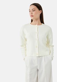 Weißer Cardigan aus weichem Stoff, mit Rundhals, langen Ärmeln und sechs Knöpfen vorne. Kombiniert mit weißen Hosen.