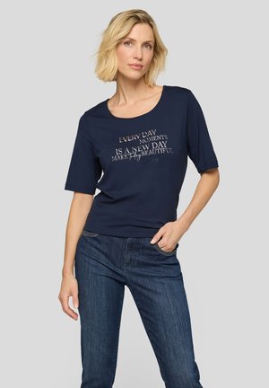 Blonde Frau in navyblauem kurzärmligen Hemd mit motivierendem Text, steht mit einer Hand in der Tasche, trägt blaue Jeans, neutraler Hintergrund.
