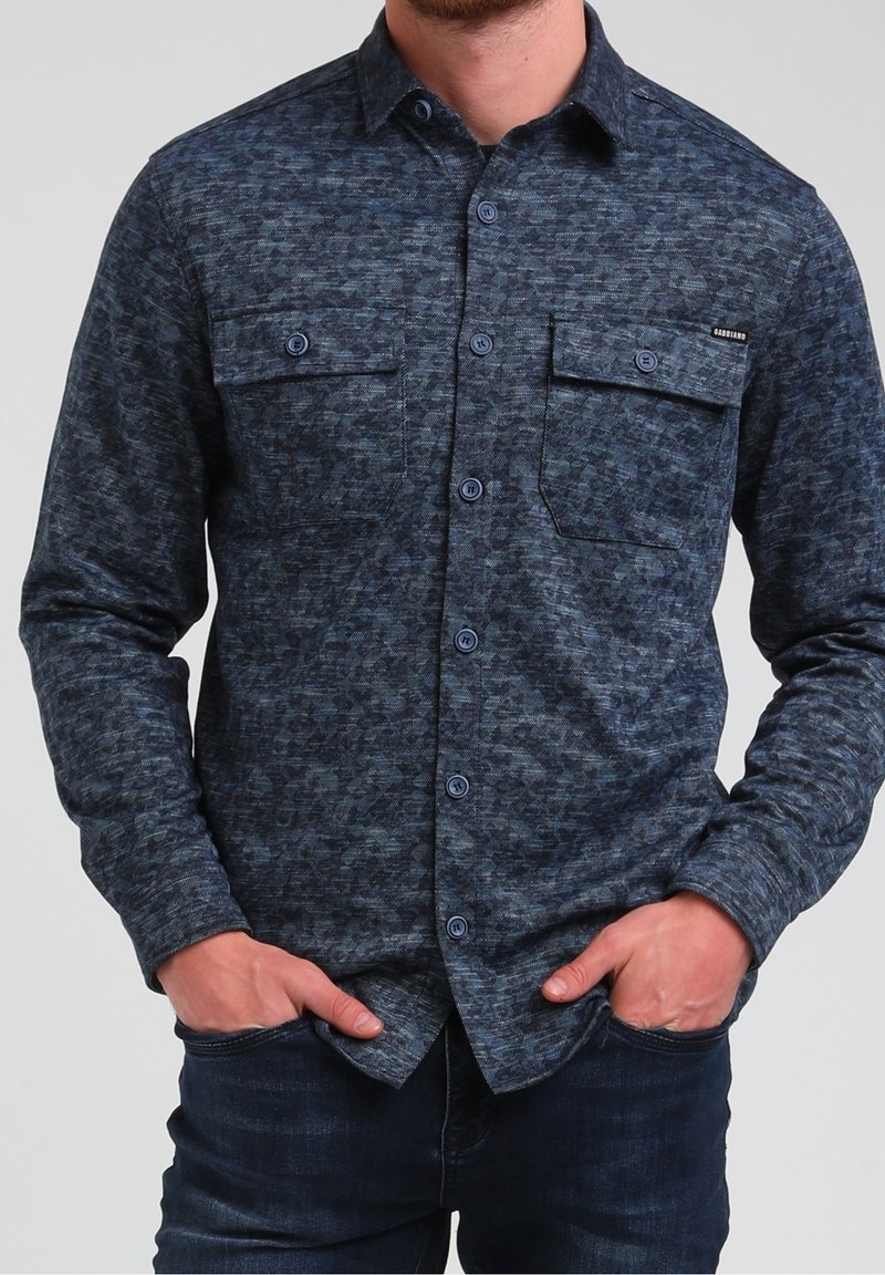 Homme portant une chemise à manches longues bleu foncé à motifs, boutonnée avec deux poches poitrine, mains dans les poches, et jean bleu foncé.