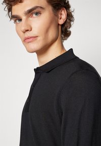 Camisola polo preta com malha texturizada, apresentando colarinho e um fecho de três botões, exibida no ombro e pescoço de um modelo.