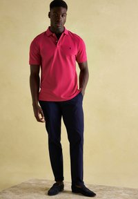 Mladý muž stojí s jednou rukou v kapse, oblečený v jasně růžovém polo tričku, tmavě modrých kalhotách a černých mokasínech na béžovém pozadí.