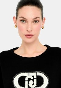 Felpa nera con un grande logo bianco impreziosito. Il modello indossa orecchini a forma di cuore in oro, con una texture liscia e lucida.