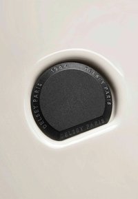 Hardware circolare nero con incisione "Delsey Paris 1946" sulla superficie di un bagaglio bianco crema, caratterizzato da una finitura liscia e lucida e bordi arrotondati.