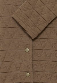 Material maro matlasat cu model de cusături triunghiulare și nasturi metalici capse pe mâneca și corpul unei haine.