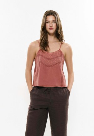 Jeune femme aux longs cheveux bruns portant un haut sans manches rose brodé de fleurs et un pantalon sombre ample, debout avec les mains dans les poches.