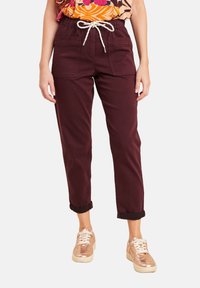 Donna che indossa pantaloni bordeaux arrotolati con tasche frontali e una cintura bianca a forma di corda, abbinati a sneakers metallizzate oro rosa.