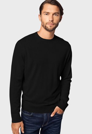 Schwarzer langärmeliger Pullover mit runder Ausschnitt und gerippten Bündchen, aus einem glatten, leichten Stoff. Kombiniert mit dunkelblauen Jeans.