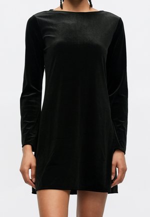 Vrouw die een zwarte velvet mini-jurk met lange mouwen en boothals draagt, staand tegen een effen lichte achtergrond.