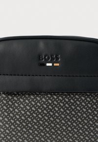 Czarna skórzana torba z teksturowanym szarym korpusem, wytłoczonym logo oraz trzema kolorowymi akcentami: czarnym, białym i brązowym w okolicy górnego szwu.