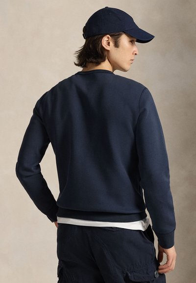 Giovane uomo di profilo che indossa un cappello blu navy, una felpa blu navy, una canottiera bianca e pantaloni cargo blu navy su uno sfondo semplice.