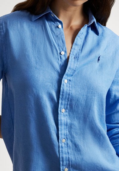 Svetlomodrá košeľa na zapínanie s dlhými rukávmi z ľanu. Má klasický golier, predný zapínací pás a malý vyšívaný logo na hrudi.