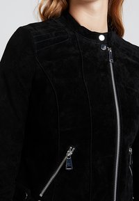 Svart mockajacka med hög krage, blixtlås framtill och snedställda sidofickor. Texturerad detaljering på axlarna ger visuell intress.