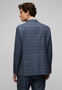 Blauwe colbert met een enkele split aan de achterkant, met gekartelde revers en knoopaccenten op de mouwen, gemaakt van een gestructureerde stof.