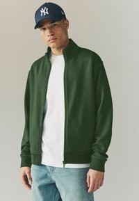 Veste zippée verte en tissu lisse, avec un col montant, des poignets côtelés, et un t-shirt blanc en dessous. Jean léger bleu clair.