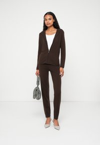 Bruine zip-up hoodie met bijpassende strakke broek, gemaakt van zachte stof. Opvallende details zijn een capuchon en slank ontwerp. Houdt een met patroon versierde tas vast.