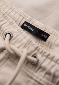 Shorts beiges avec une taille à cordon de serrage et des œillets métalliques, ornés d'une étiquette noire "ombre" ; tissu doux avec une texture subtile.