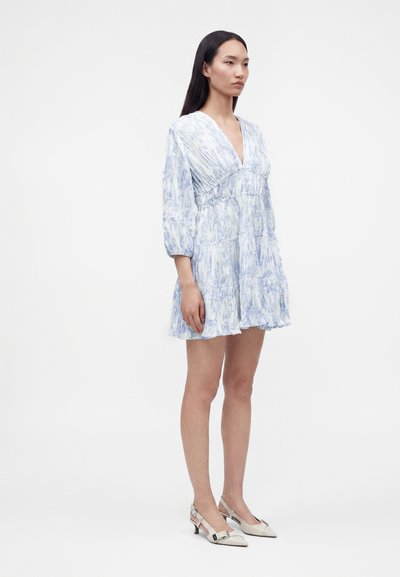 Femme aux longs cheveux foncés porte une robe bleu clair à motifs avec un col en V, des manches trois-quarts et des chaussures slingback beige à petit talon, debout de côté.