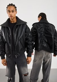 To personer med flettehår iført svarte skinnjakker og grå jeans, en som vender fremover og den andre som viser baksiden av jakken.