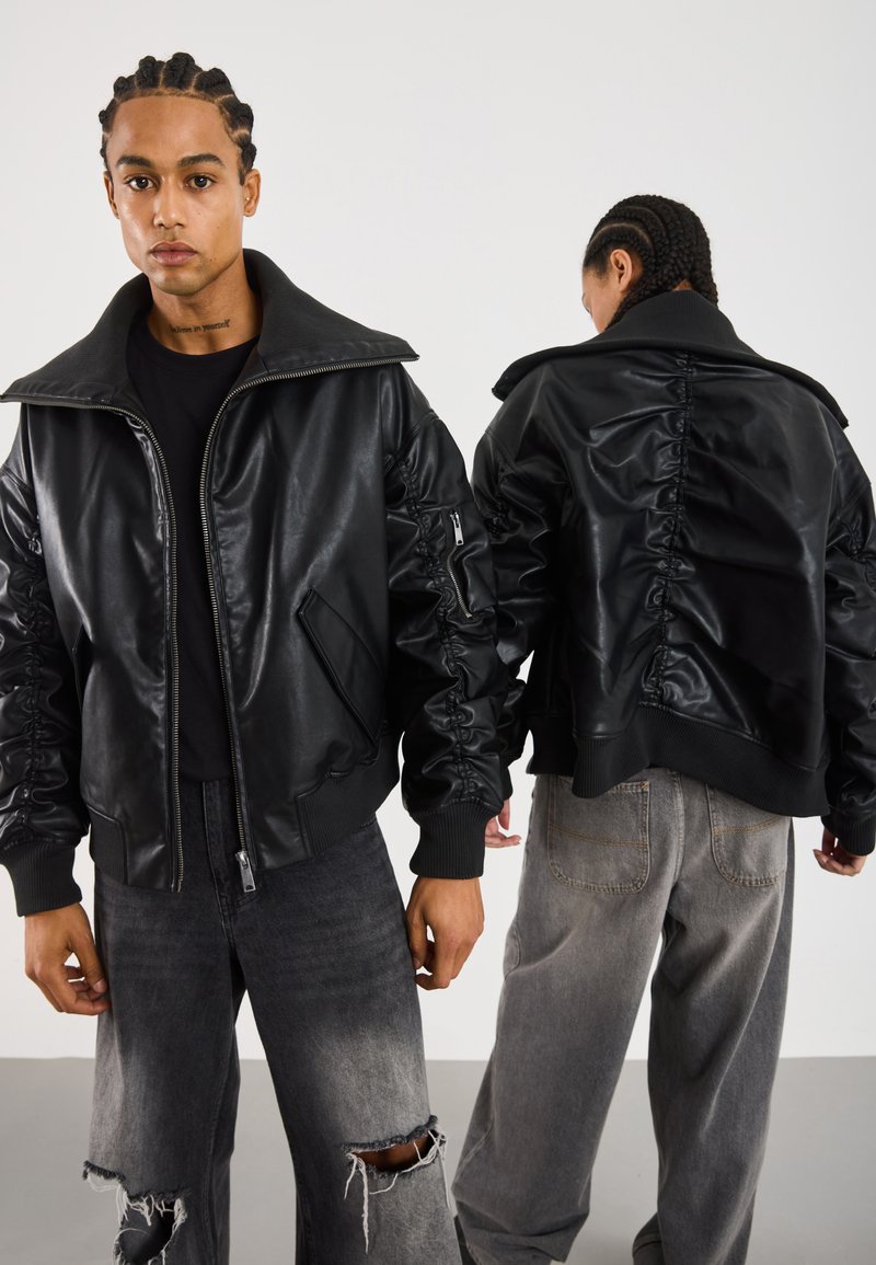 Due persone con capelli intrecciati, indossano giacche di pelle nera e jeans grigi, una di fronte e l'altra che mostra il retro della giacca.