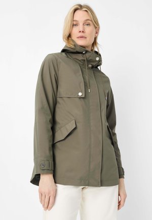 Frau trägt eine olivgrüne Kapuzenjacke mit vorderen Klappentaschen und Knopfdetails, kombiniert mit weißen Hosen, vor einem schlichten Hintergrund.