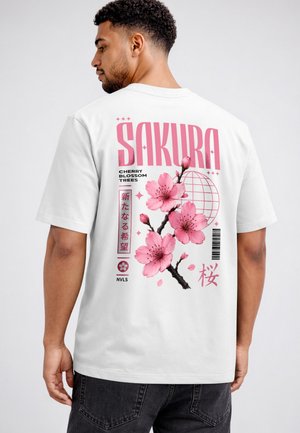 Man wearing white t-shirt with large pink cherry blossom branch, globe graphic, and "SAKURA" text on the back.