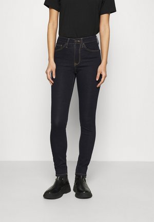 Jeans skinny bleu foncé en tissu denim avec des coutures visibles, taille haute, conception à cinq poches et fermeture à bouton à l'avant.