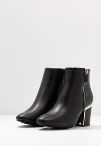 Botines de cuero negro hasta el tobillo con punta afilada, cremallera lateral y un tacón grueso con detalles en dorado. Textura suave.