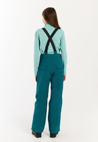Tyrkysové snehové nohavice s čiernymi traky a bočnými vreckami, v kombinácii s svetlomodrým dlhým rukávom. Hladká, odolná tkanina, uvoľnený strih.