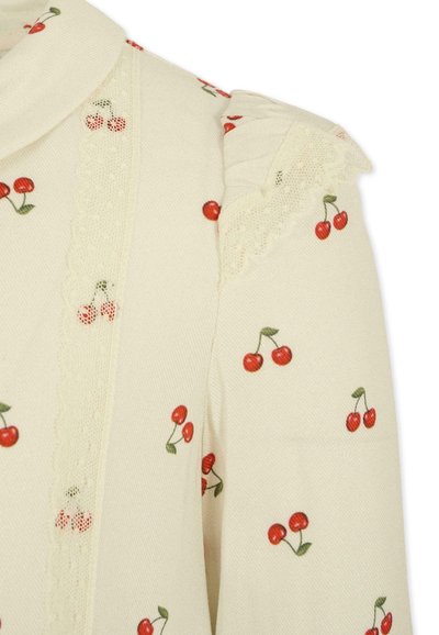 Blusa di tessuto color crema con stampa di ciliegie. Presenta un bordo in pizzo lungo la parte anteriore e dettagli arruffati sulla spalla.