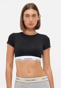 Svart avkortet t-skjorte laget av myk stoff, med korte ermer og et hvitt belte med et gjentakende Calvin Klein-logo.