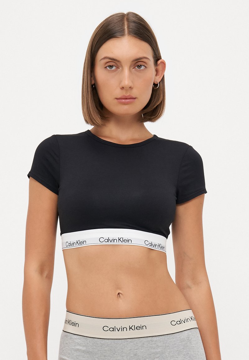 Svart avkortet t-skjorte laget av myk stoff, med korte ermer og et hvitt belte med et gjentakende Calvin Klein-logo.