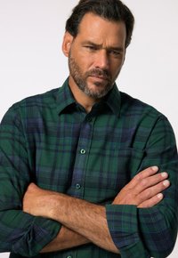 Camisa de franela a cuadros verdes y azul marino con mangas largas, cuello abotonado y un bolsillo en el pecho. Presenta botones verdes y una textura suave.