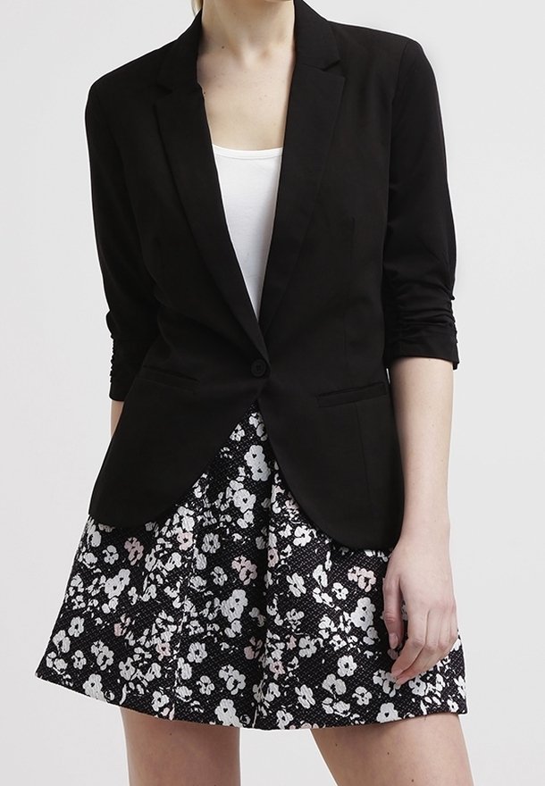 Femme portant un blazer noir avec des manches froncées sur un top blanc, associé à une jupe noire avec un motif floral blanc et rose clair.