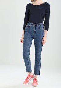 Žena v těsném tmavě modrém topu s dlouhým rukávem, v výrazně modrých džínách se zvýšeným pasem a s roztrhaným lemem, a v růžových sportovních teniskách s bílými pruhy.