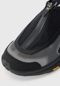 Chaussure de sport noire avec une entrée zippée, tige en mesh et textile ornée de motifs en pointillé, et semelle en caoutchouc texturé marquée "Vibram."
