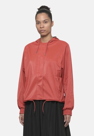 Mujer con chaqueta roja con capucha, cordones y puños elásticos, combinada con una falda negra, de pie frente a un fondo liso.