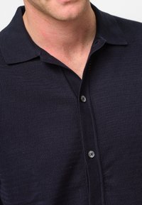 Polo shirt navy a maniche corte con tessuto testurizzato, colletto piatto e una patta con cinque bottoni. I bottoni sono scuri e di forma circolare.