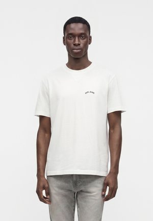 Mies, jolla on yllä yksivärinen valkoinen Pepe Jeans -t-paita ja vaaleanharmaat farkut, seisoo valkoista taustaa vasten, kasvot kohti katsojaa.